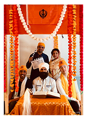 Photo of 4 men and a woman in traditional indian attire, surrounded by an orange fabric altar with white, yellow and deep orage garlands. A white man with a beard sitting at the center of the group.