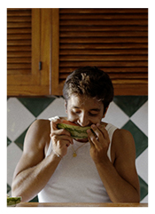 Man with dark brown hair, wearing a tank-top and eating a water Mellon. behind him are light brown wooden kitchen cabinet doors on top. White and green checkered tiles below.
