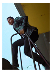 Man with black pants and long-sleeved dark metallic green jacket, holding onto a rail. Yellow ceiling above him.